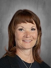 A smiling woman with shoulder-length brown hair and light makeup, wearing a black sweater, against a gray background.