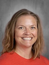 Smiling woman with medium-length brown hair, wearing an orange shirt, set against a gray background.