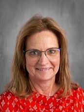 Woman with glasses and shoulder-length hair, wearing a red patterned top, smiles against a gray background.