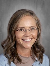Smiling woman with long, wavy brown hair and glasses, wearing a light blue cardigan against a gray background.