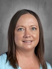 Headshot of a woman with long brown hair, smiling, wearing a light blue shirt against a gray background.