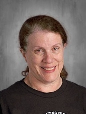 A person with brown hair tied back, wearing a black shirt, smiling against a gray background.