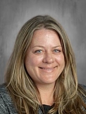 Headshot of a smiling woman with long, wavy hair, wearing a black shirt and gray jacket, set against a neutral background.