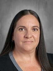 A woman with long dark hair and a neutral expression poses for a portrait against a gray background.