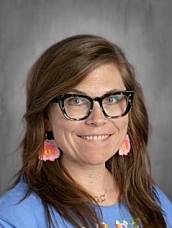 A smiling woman with long brown hair, wearing glasses and colorful earrings, against a gray background.