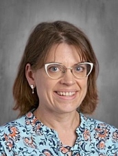 A woman with brown hair and glasses smiles, wearing a floral blue blouse, against a gray background.