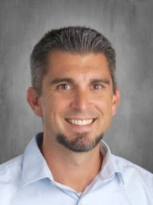 Smiling man with short gray hair and a goatee, wearing a light blue shirt against a gray background.